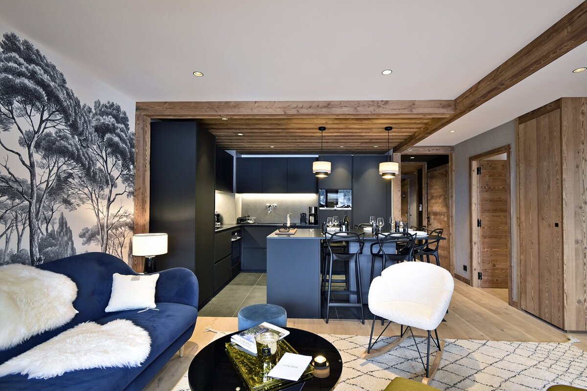 Modern open-plan living room and kitchen/dining area with a blue sofa, black cabinetry, wooden beams, and a tree mural accent wall.