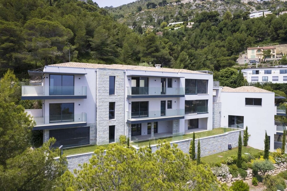 A modern multi-story building with glass balconies and terraced gardens, nestled on a lush, forested hillside with other homes.