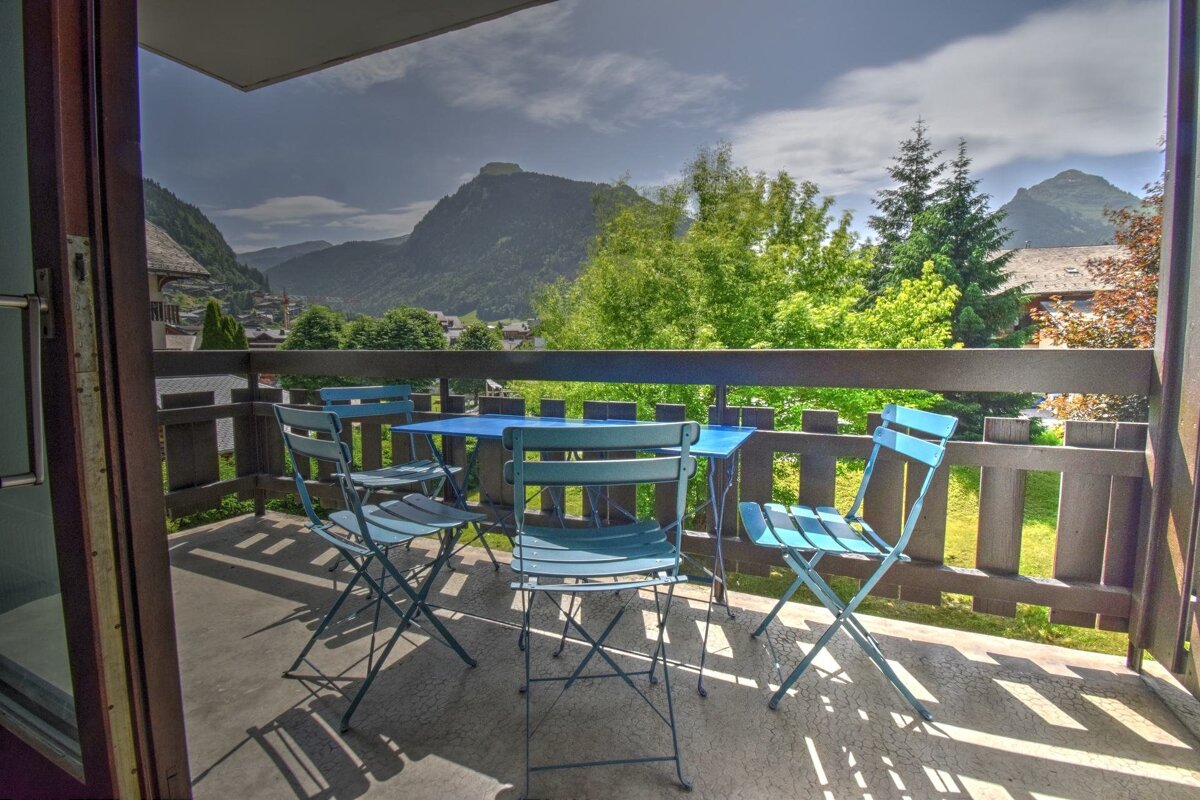 A balcony with blue table and chairs overlooks a scenic mountain village nestled among green trees and hills under a partly cloudy sky.