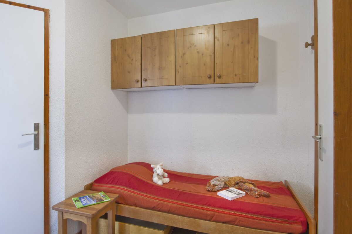A small, cozy room with a single bed featuring red and orange striped bedding, a plush toy, and a book. Wooden cabinets are mounted above the bed.