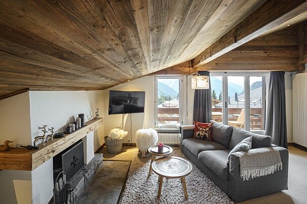 Cozy chalet living room with a slanted wood ceiling, fireplace, grey sofa, and large windows looking out to mountain scenery.
