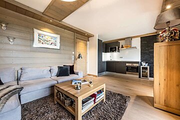 Cozy open-plan living room with wood-paneled walls, grey sofa, wooden coffee table, and a modern kitchen featuring dark cabinets and a black stone accent wall.