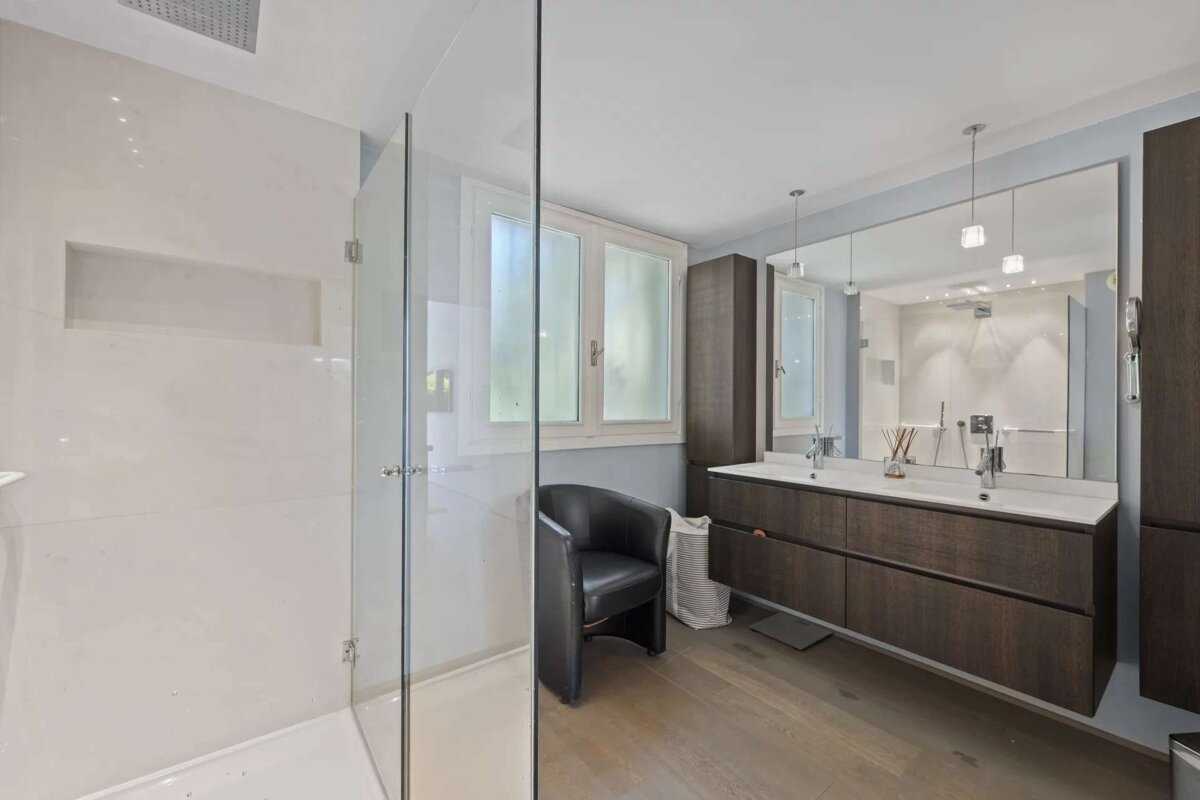 Modern bathroom featuring a glass shower, double dark wood vanity with a large mirror, light wood floor, window, and a black armchair.