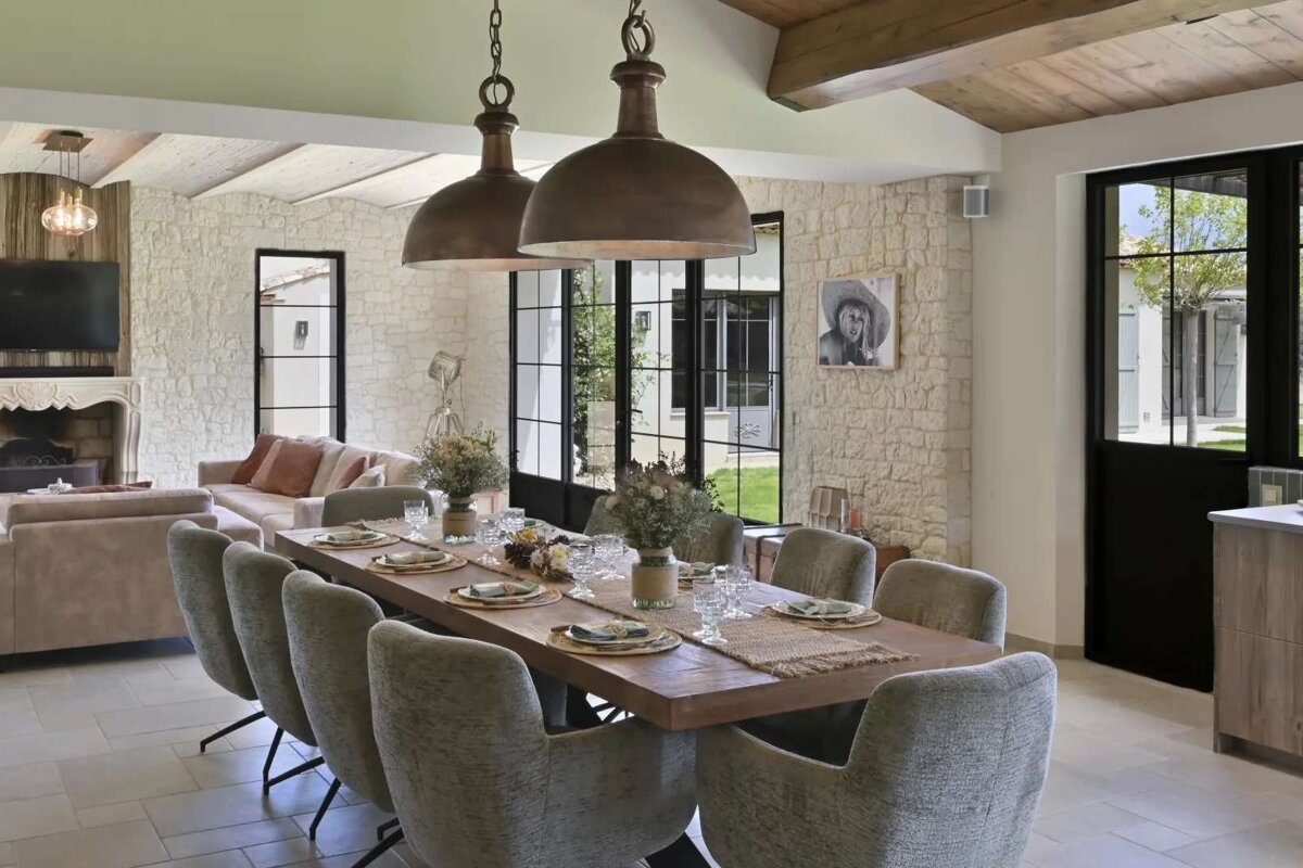 Spacious, rustic-chic dining and living area with stone walls, a long set table, cozy sofas, and large industrial-style windows opening to an outdoor view.
