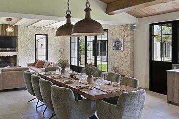 Spacious, rustic-chic dining and living area with stone walls, a long set table, cozy sofas, and large industrial-style windows opening to an outdoor view.