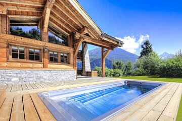 A wooden chalet with an outdoor plunge pool built into a deck, set against a stunning mountain and forest landscape under a bright blue sky.