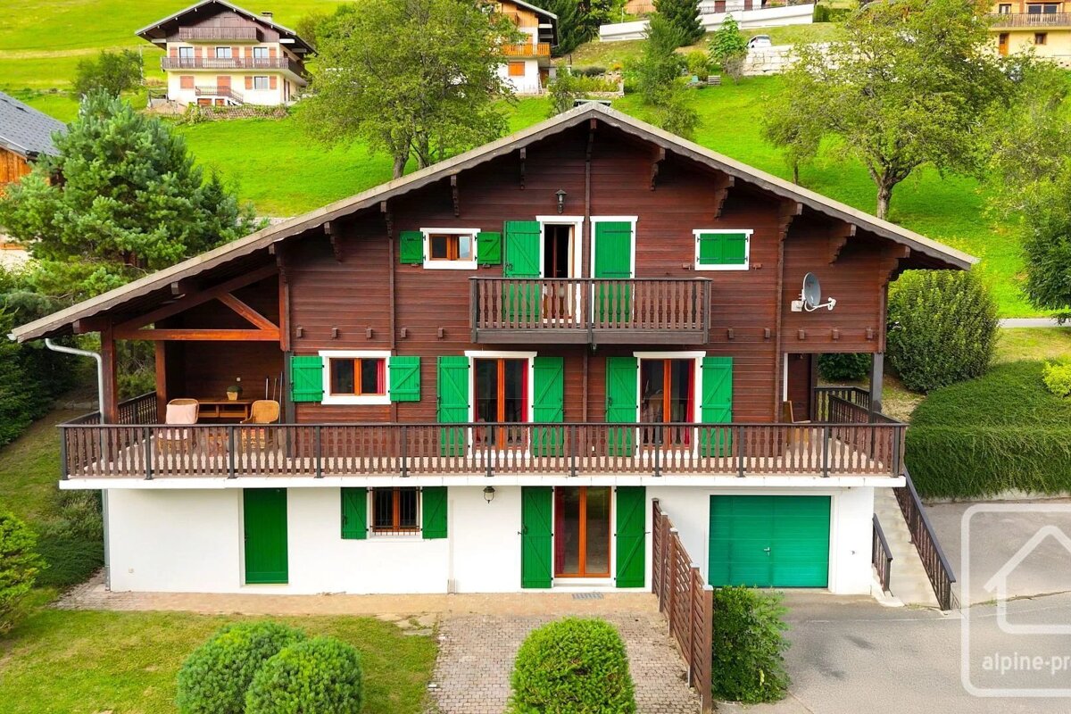 A wooden chalet-style house with bright green shutters and balconies, set against a lush green hillside.