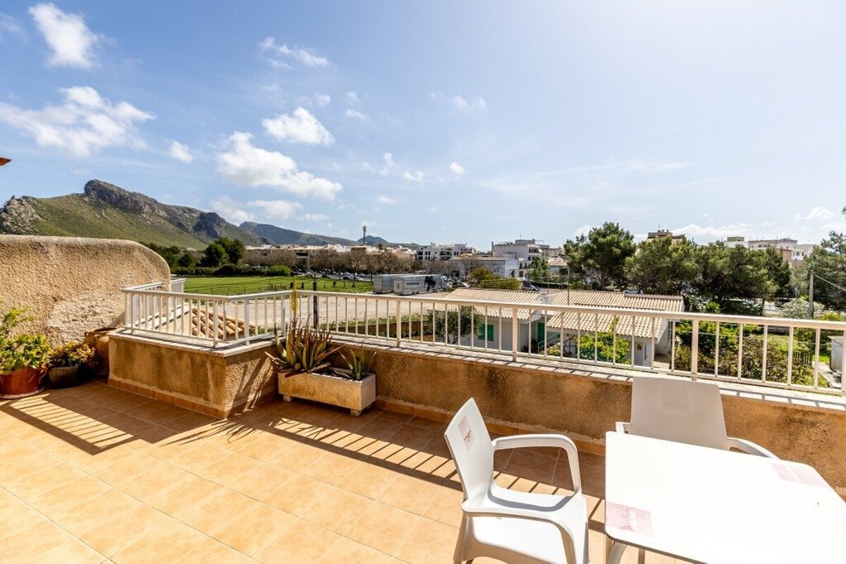 Sunny tiled balcony with white table and chairs, offering views of a town, green fields, and distant mountains under a blue sky.