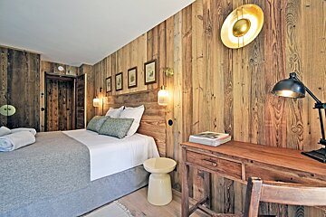 A warm, rustic bedroom with full wood paneling, a bed with soft linens, and a wooden desk under a modern lamp.
