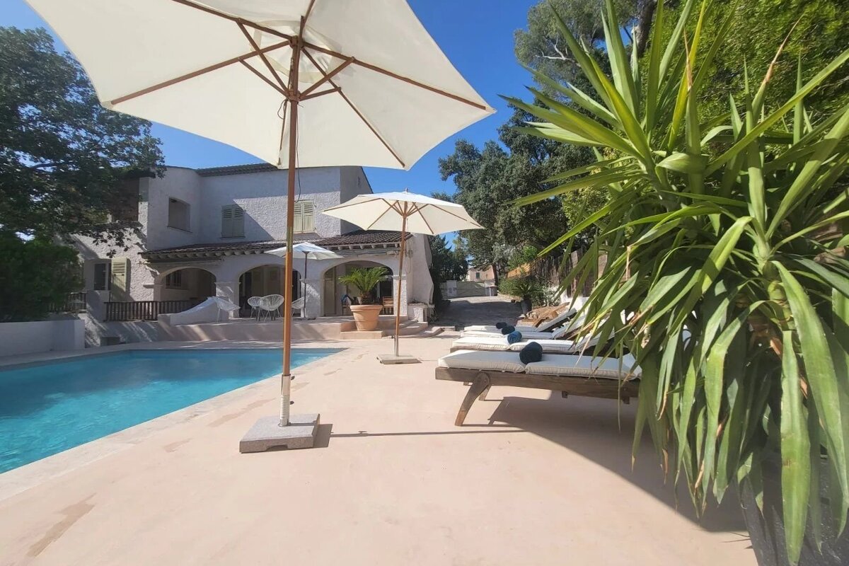 A sunny poolside scene with a villa, umbrellas, sun loungers, and lush plants under a clear blue sky.