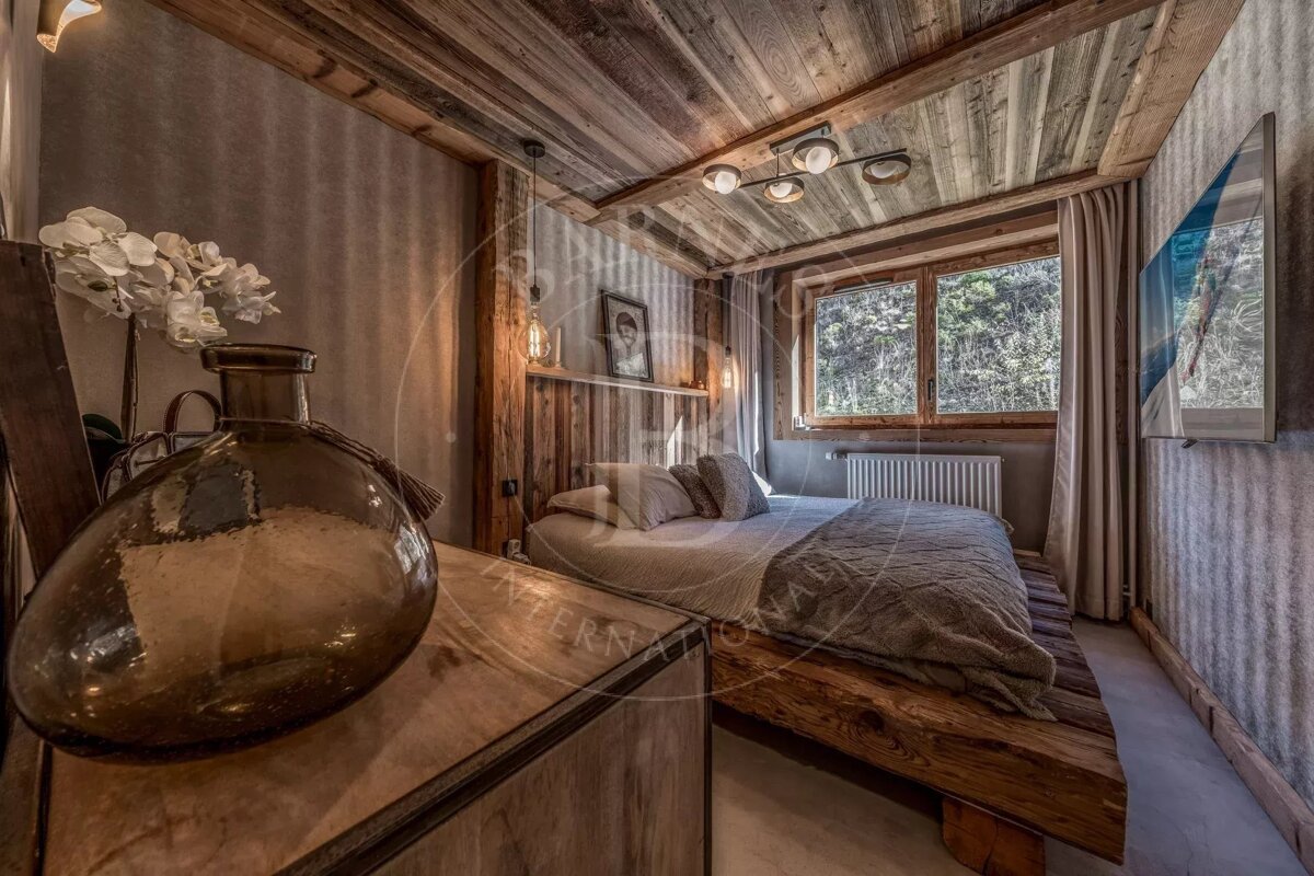 A cozy, rustic bedroom featuring warm wooden walls/ceiling, a large bed, and a window with a natural view. A modern TV and chic decor complete the space.