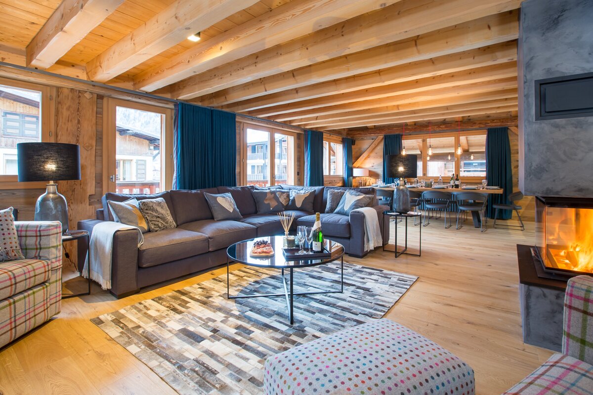 A spacious, modern rustic living room with a large gray sectional, wooden ceiling beams, a cozy fireplace, and a dining area, accented by blue curtains.