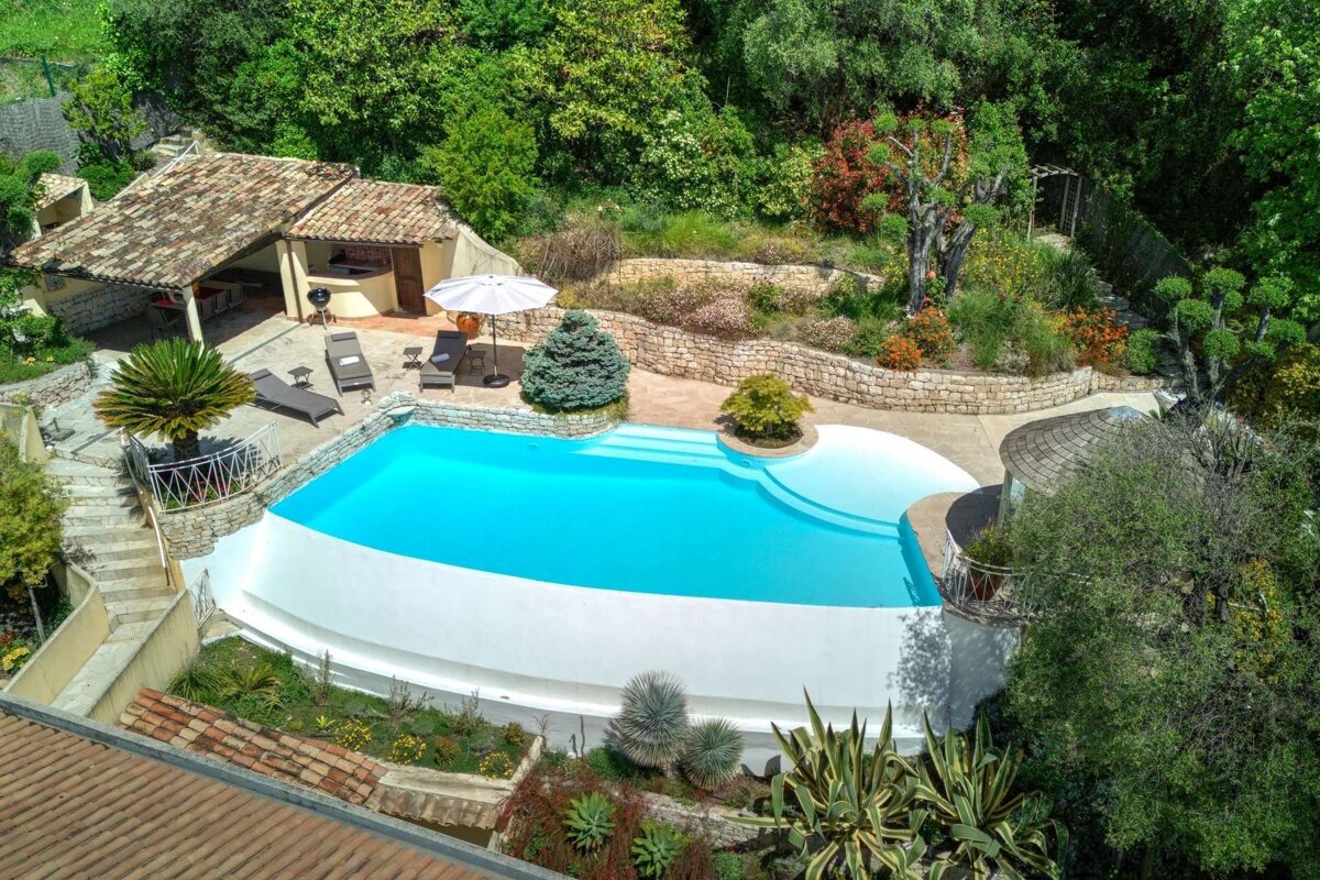 Aerial view of a vibrant blue pool, stone patio, outdoor kitchen, and lush terraced gardens surrounded by green trees.