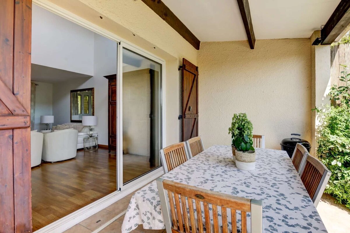 A covered patio features a dining table with chairs and a plant, connected by a sliding glass door to an indoor living room with a sofa. Wooden shutters and a grill are visible.