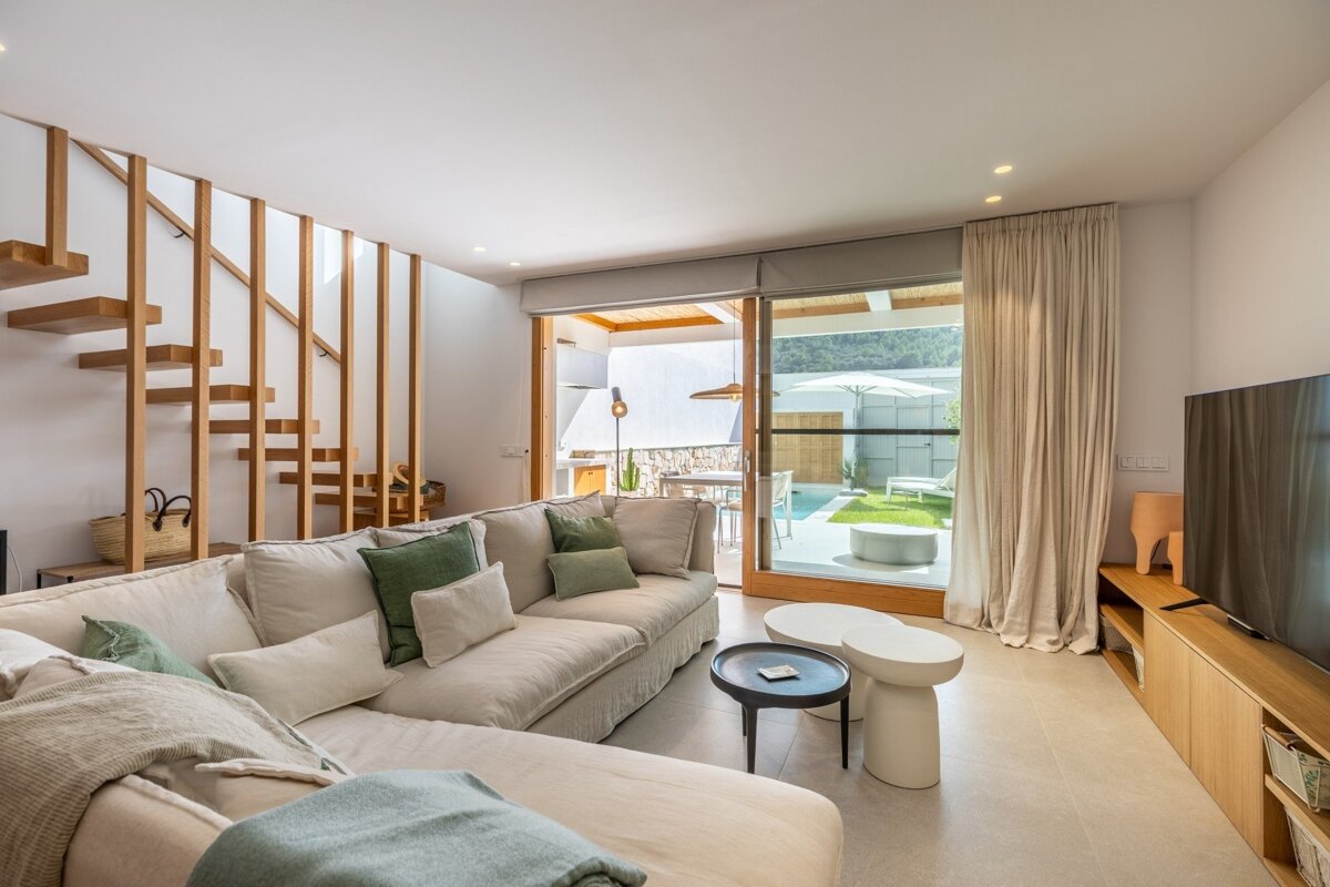 Bright modern living room with a large sofa, wooden floating stairs, and glass doors opening to a patio with a pool and green space.
