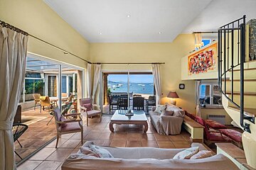 A spacious, sunlit living room featuring cozy seating, tiled floors, and sliding glass doors that open to a scenic view of the sea.