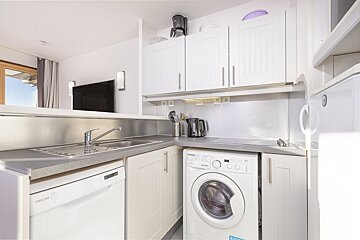 A modern white kitchen with integrated dishwasher, washing machine, sink, and upper cabinets. An open layout connects to a living area with a TV.