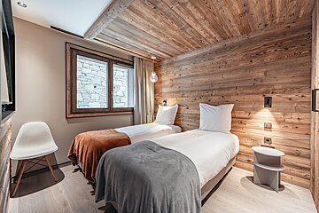 A cozy, wood-paneled bedroom with two twin beds, one with a grey throw and one with an orange throw, a white accent chair, and a window.