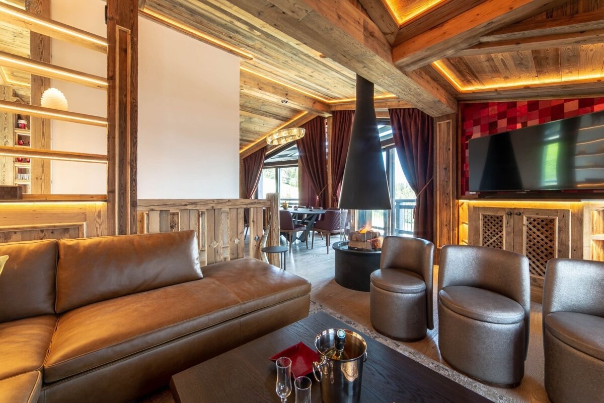 A luxurious chalet living room features rich wood, a modern hanging fireplace, a large leather sofa, and a dining area. Champagne awaits on the coffee table.