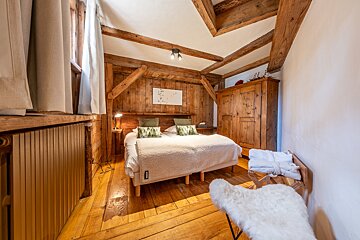 A cozy, rustic bedroom with warm wooden walls, floor, and exposed beams. Features a double bed, wooden wardrobe, and a fluffy white chair for an inviting retreat.