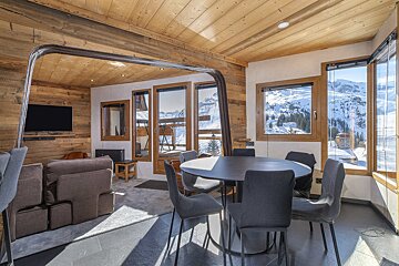 Cozy wooden cabin interior featuring a dining table, living area, and large windows with a scenic view of snowy mountains.