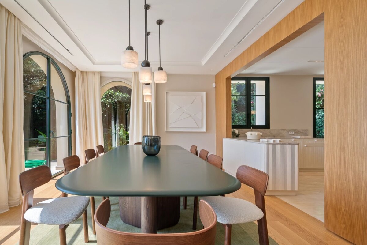 Elegant dining room featuring a green oval table, wooden chairs, pendant lights, and large arched doors opening to a garden.