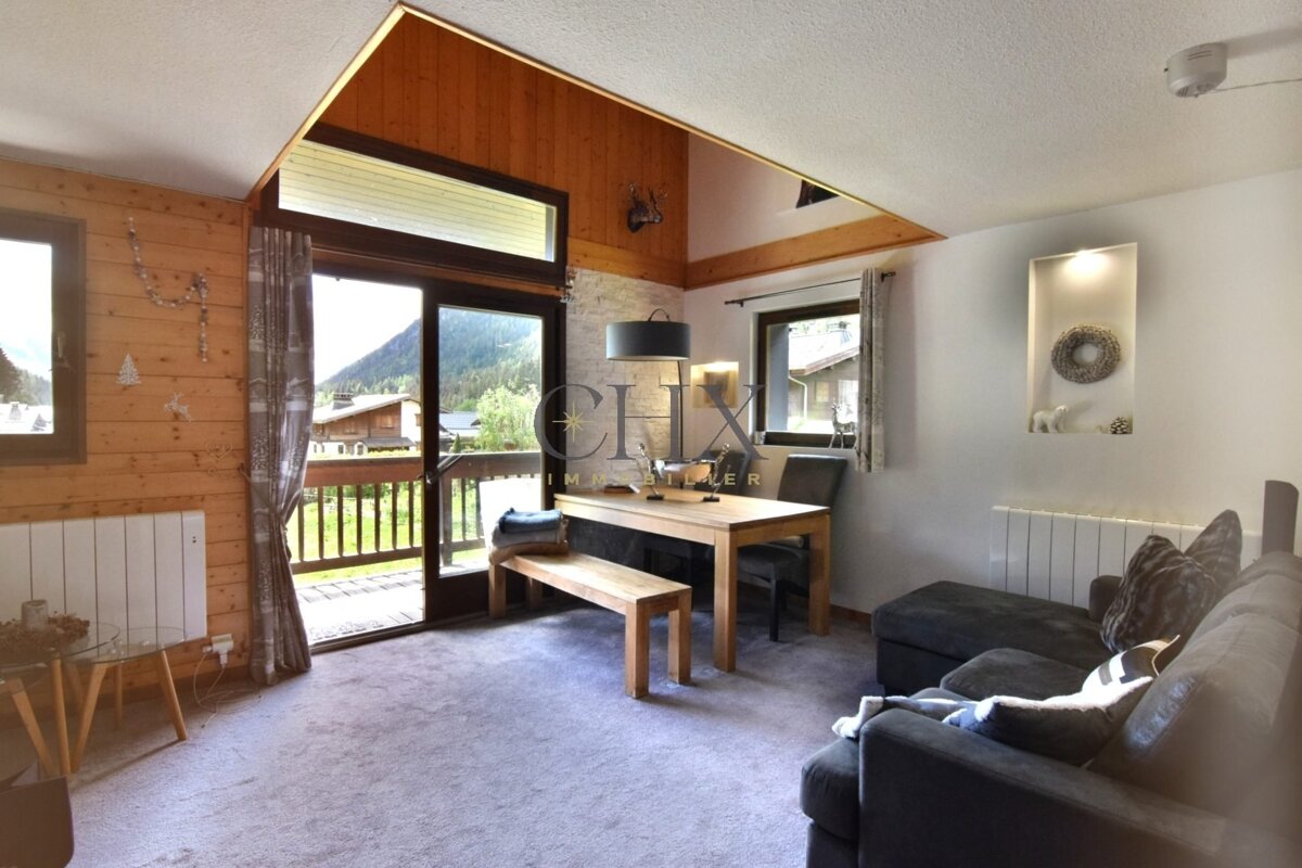 Cozy alpine chalet interior with wooden paneling, dining table, grey sofa, and sliding glass doors showing mountain scenery.