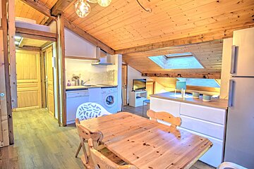 Bright wooden attic apartment with a dining table, kitchenette, white cabinets, and sloped wood ceilings with skylights.