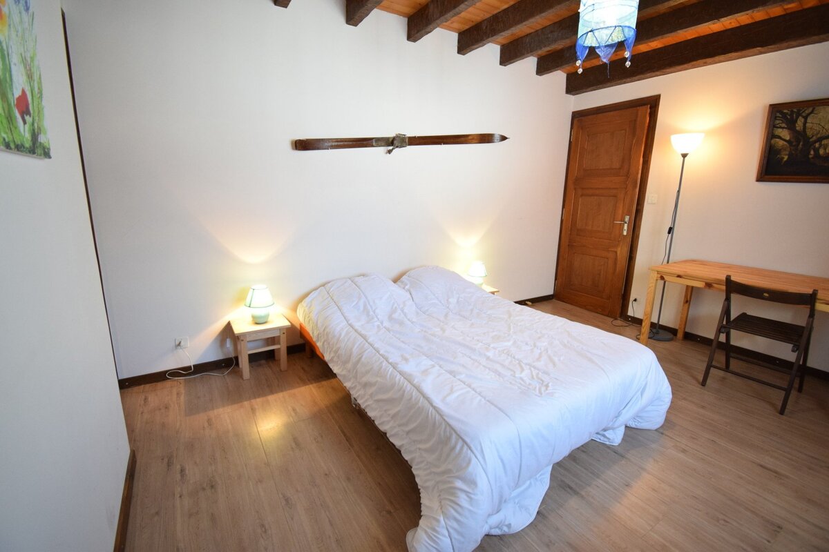 A cozy bedroom with white walls, wooden floor, exposed beams, a bed with white duvet, bedside lamps, vintage ski decor, desk, and a blue ceiling lamp.