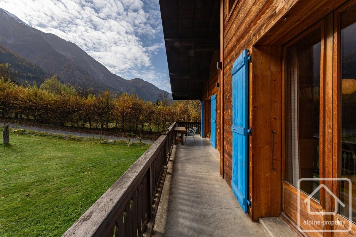 A wooden chalet balcony with blue shutters overlooks a green field, autumn trees, and majestic mountains under a cloudy sky.