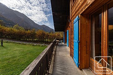 A wooden chalet balcony with blue shutters overlooks a green field, autumn trees, and majestic mountains under a cloudy sky.