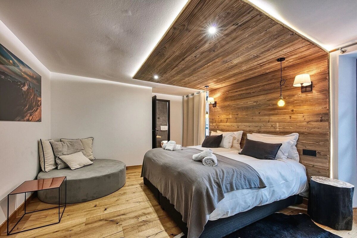 Stylish bedroom with warm wood paneling on the headboard and ceiling, modern accent lighting, a large bed, and a round chaise lounge.