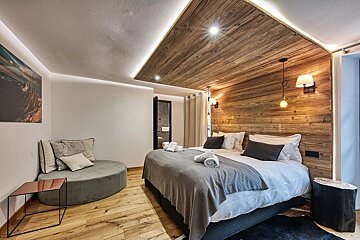 A modern, cozy bedroom with a wooden accent wall/ceiling, large bed, and round grey lounge. Warm lighting and chic decor.