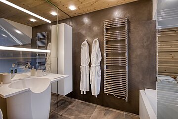 Sleek modern bathroom with a white sculpted sink, dark textured walls, wooden ceiling, heated towel rack, and two cozy robes.