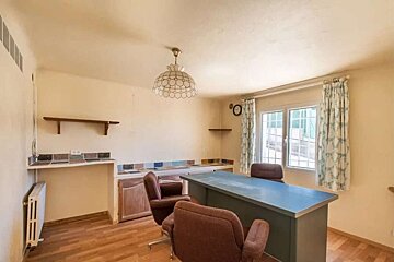 Neutral-toned room with wood floors, a large blue-topped table, brown office chairs, wall shelves, and a window with patterned curtains.