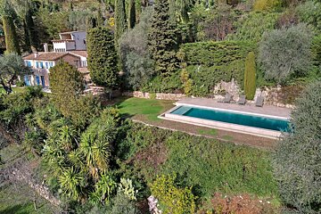Aerial view of a charming house with blue shutters, a long swimming pool, and verdant Mediterranean vegetation on a hillside.
