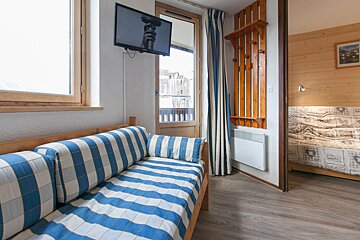 A cozy room features a blue and white striped daybed, a wall-mounted TV, balcony door, and a wooden coat rack. An adjacent bedroom is also visible.