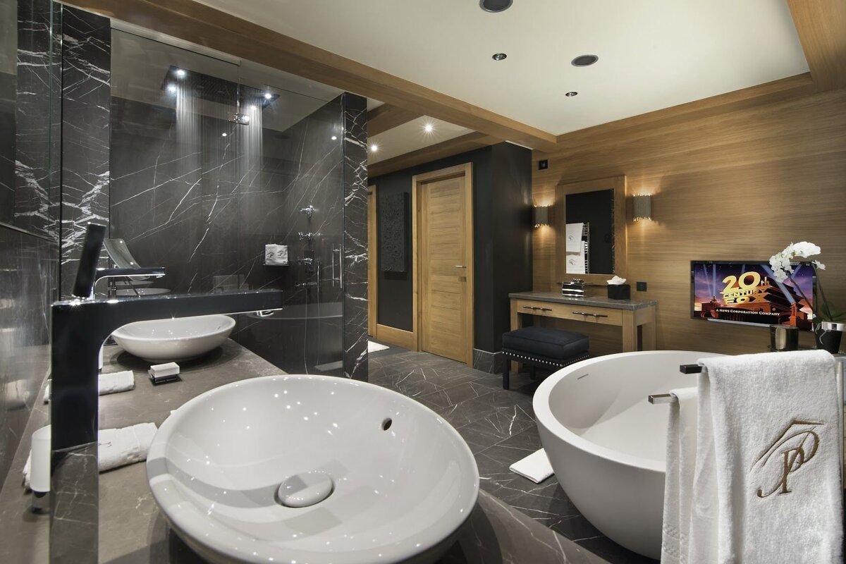 Luxurious modern bathroom featuring black marble and wood decor. It includes a rain shower, dual sinks, freestanding tub, and a TV showing 