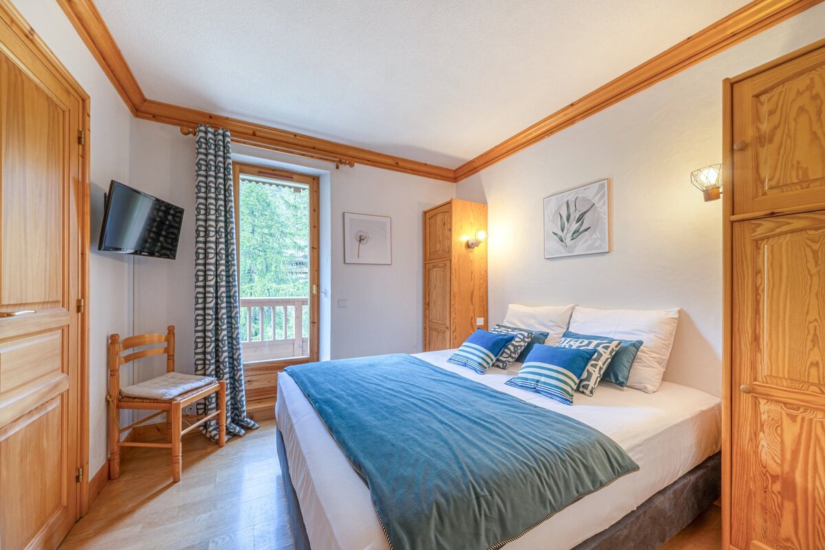 Cozy bedroom with wooden features, a large bed with blue bedding, wall-mounted TV, and a balcony view of green trees.