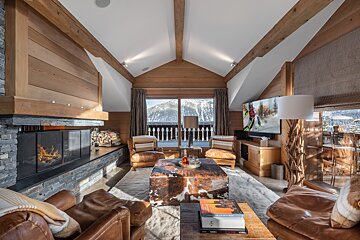 Cozy alpine chalet living room with a stone fireplace, leather armchairs, and large windows overlooking snowy mountains.