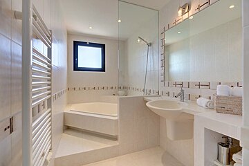 A bright, modern white bathroom features a built-in tub, walk-in shower with glass panel, pedestal sink, large mirror, towel warmer, and window.