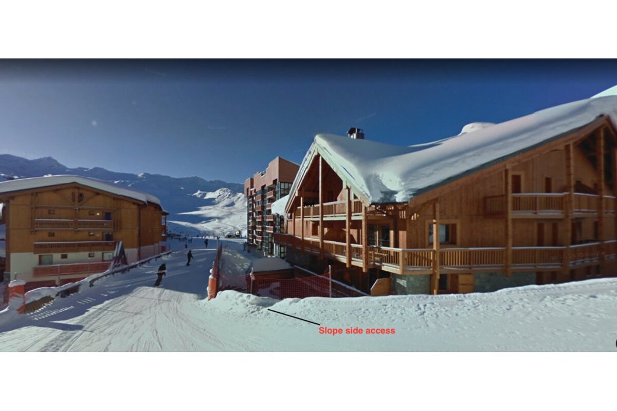 A ski resort with snow-covered wooden chalets, skiers on a path, and red text indicating "Slope side access" on the snow.