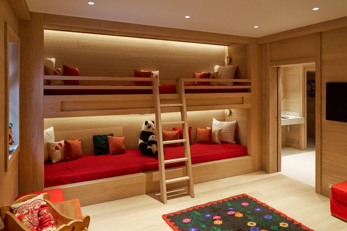 A cozy wooden kids' room with built-in bunk beds, red bedding, warm lighting, panda plushies, and a colorful floral rug.