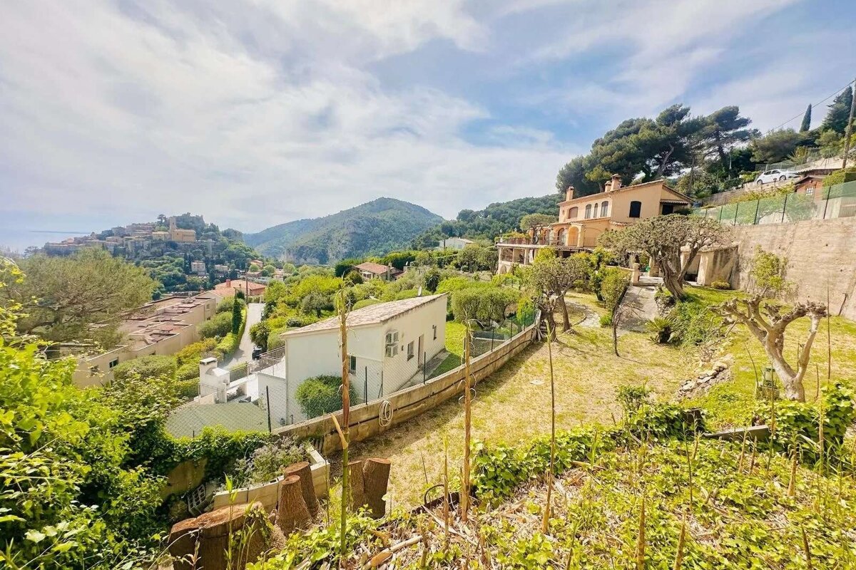Hillside homes amidst lush greenery, overlooking a distant fortified village and mountains under a cloudy sky.