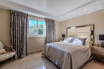 A modern, neutral-toned bedroom with a large bed, patterned curtains, a window revealing trees, and reflective tiled flooring.