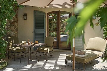 A sun-dappled outdoor patio with a dining set, chaise lounge, and umbrella. French doors and blue shutters are visible on the building facade.