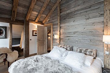 Cozy, rustic bedroom featuring exposed wooden beams, distressed wood walls, a bed with furry blankets/pillows, and a fluffy chair, evoking a chalet feel.