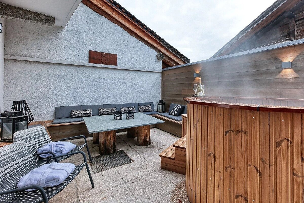 A cozy outdoor patio featuring a steaming wooden hot tub with champagne, and a seating area with a table and chairs.
