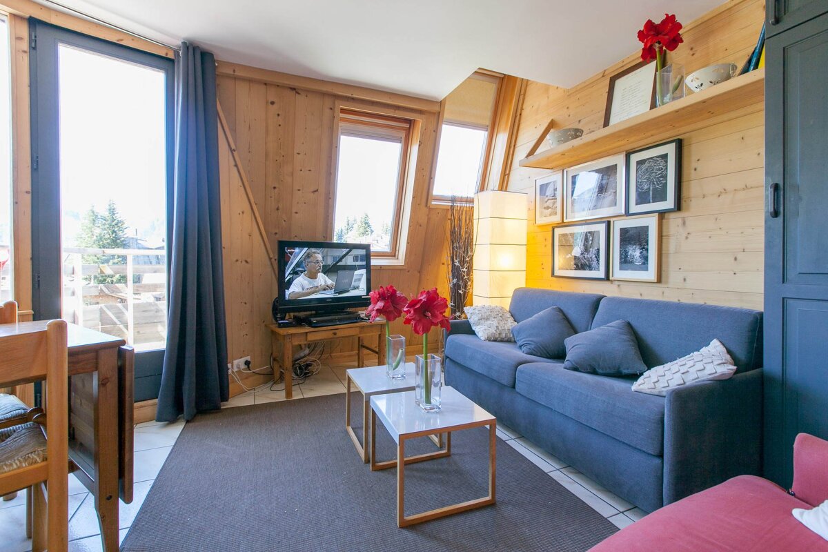 Bright, wood-paneled chalet living room featuring a blue sofa, TV, red flowers, and large windows leading to a balcony.