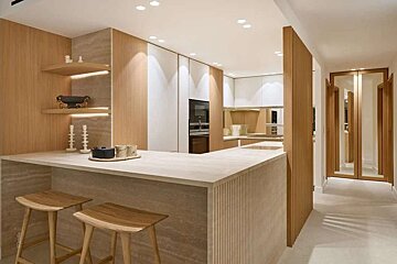 A chic, minimalist kitchen features light wood, white cabinets, a stone bar with stools, and integrated lighting. A hallway with mirrored doors is visible.